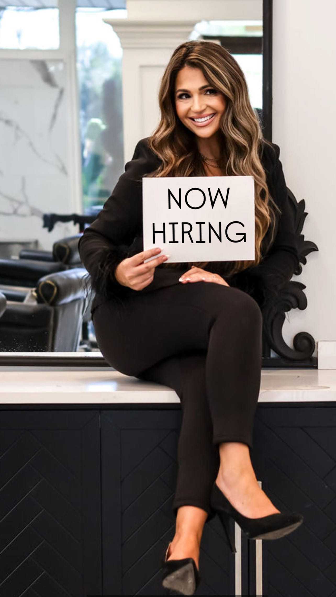 Woman in black outfit holding a "Now Hiring" sign, sitting in a stylish interior.